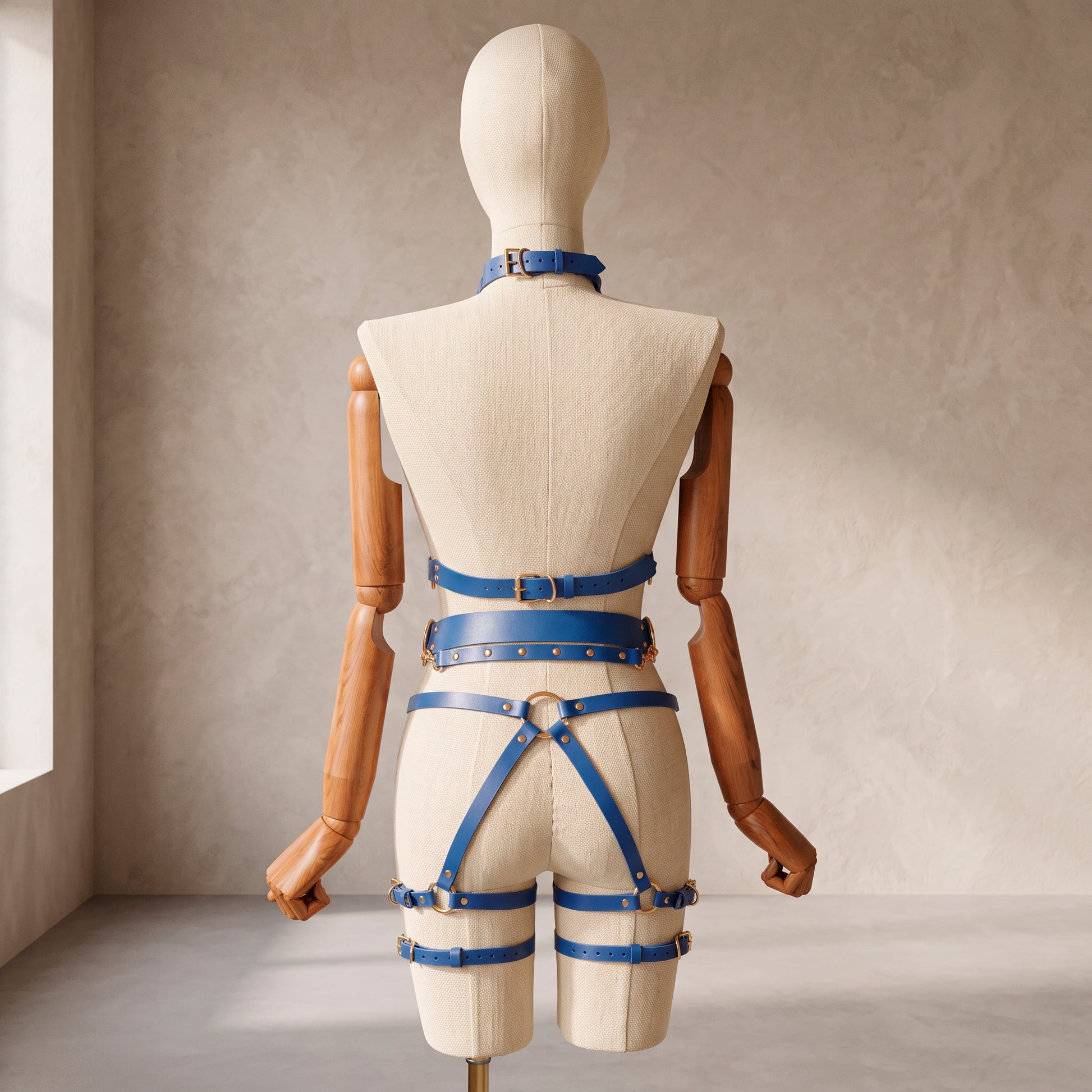 Back view of a deep blue leather full body  harness set with an open bust harness, open crotch garter belt, removable leather handle, large front ring, adjustable straps, and gold-tone metal hardware.