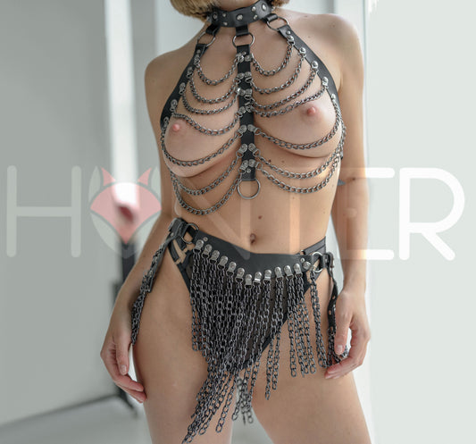 Front view of a black leather festival set with dark nickel hardware, showing a chain drape chest harness with a high collar, layered suspended chains across the chest, and a chain skirt belt with a large front chain panel and side draped chain sections.