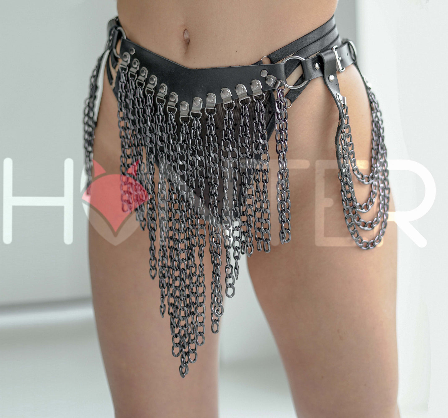 Front angled view of black leather chain skirt harness worn on a model. Multiple dark nickel chains cascade in a triangular formation at the front, with curved side chains draping along the hips. Adjustable buckle straps are visible on both sides.