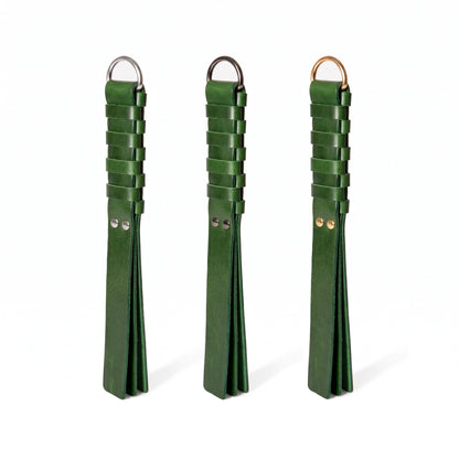 Three dark green leather flogger-style accessories with woven strap sections and metal rings, displayed side by side on a white background. Hardware in Silver, Dark nickel and gold colors.