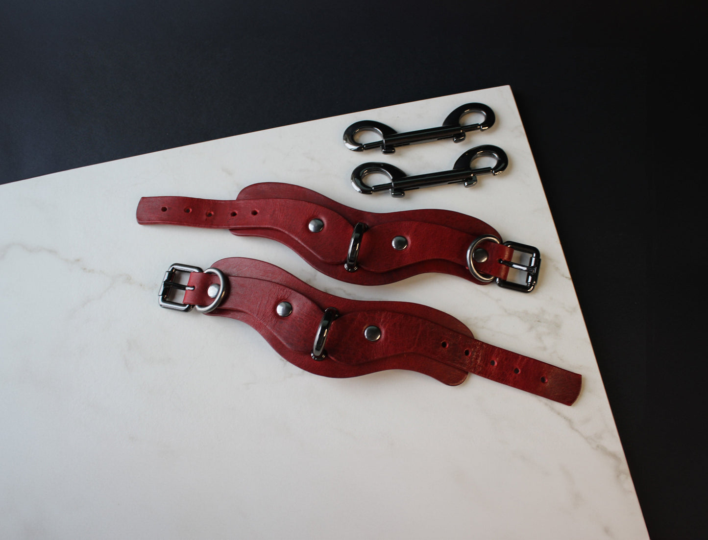 Two red leather handcuffs with metal buckles on a marble surface.