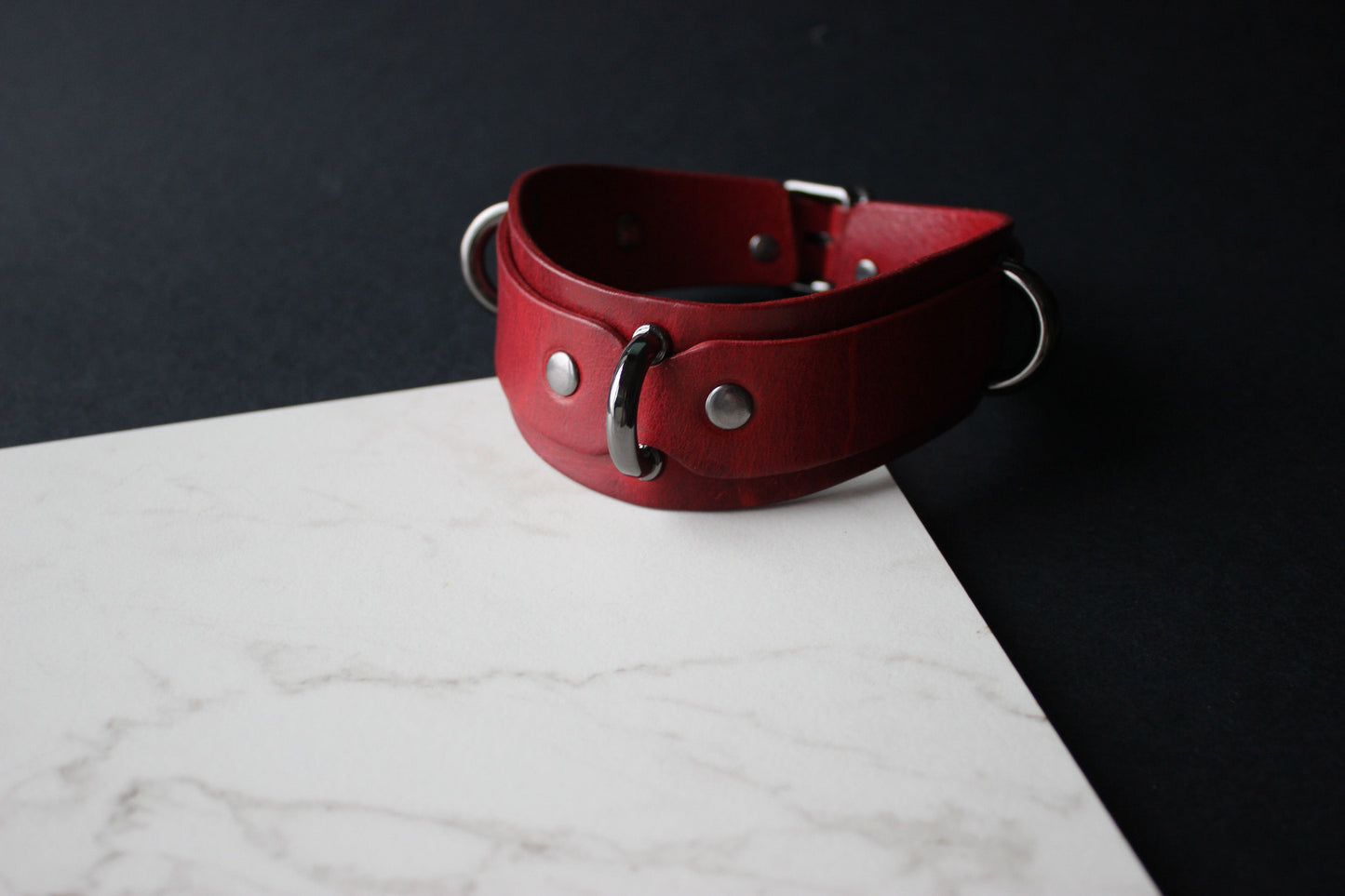 Red leather collar with silver hardware on a marble surface with a black background